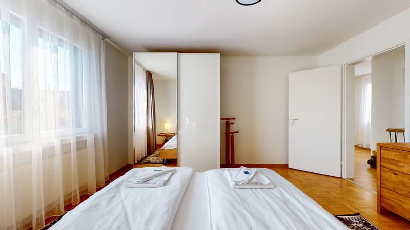 Bedroom with a double bed, large windows, white curtains, a wooden floor, white and wooden wardrobe, a nightstand with a lamp, and another bedroom with a single bed visible through the doorway.