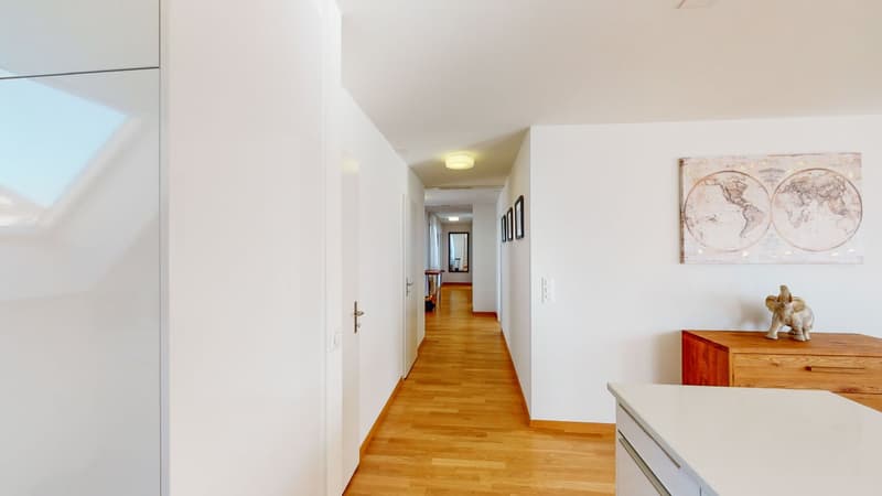 hallway with wooden floor and white walls, lit by ceiling lights, leading to a room