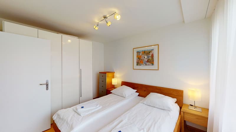 Bedroom with two single beds, wooden headboard, white linens, two nightstands with lamps, wooden cabinet with drawers, mounted artwork, and white doors and curtains