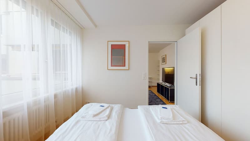 modern room, white walls, white curtains, picture frames, white bed with towels, white wardrobes, open door to another room with TV and shelves