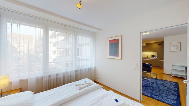 Bedroom with two beds, large window with curtains, framed picture on the wall, hardwood floors, lamp, hallway with kitchen, rug, shelf, and chair.