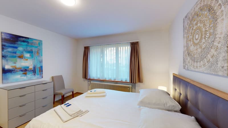 large bed, bedside table, dresser, chair, large paintings, window with white curtains