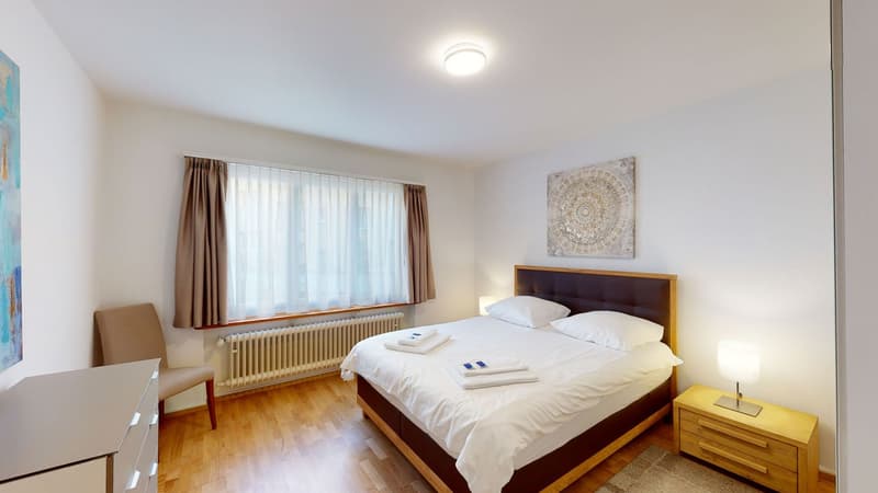 Bedroom with a large bed, wooden floor, white walls, beige armchair, white nightstand with lamp, white headboard, radiator, large window with curtains, artwork on the wall