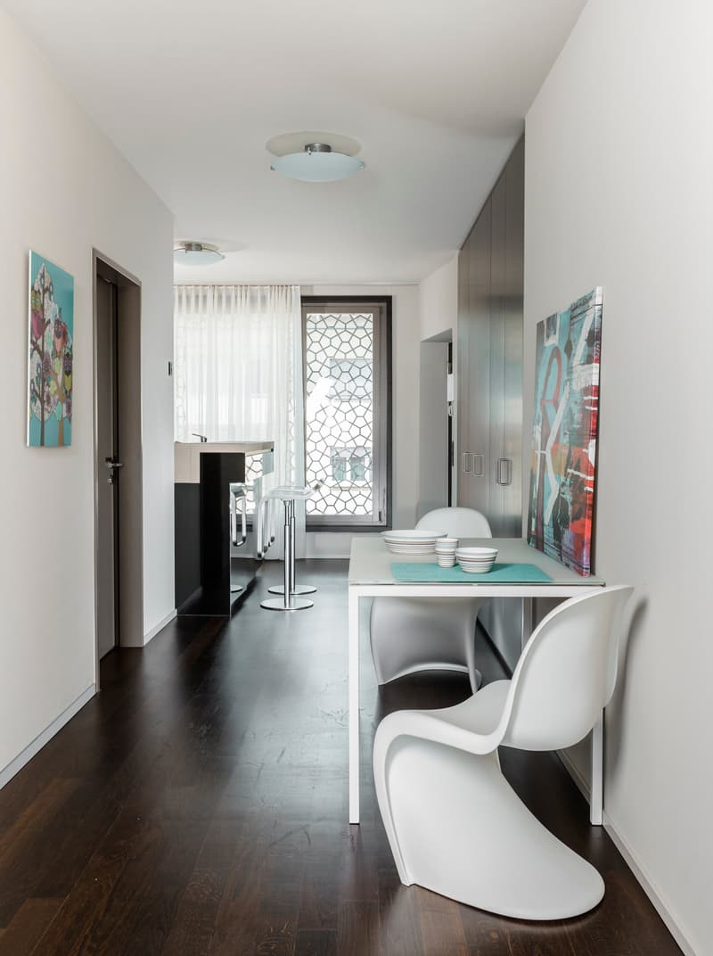 A contemporary dining area with white walls, wooden floor, and modern furnishings. Includes a white table, two white chairs, a counter with stools, a glass door, and a glass window. Decor features a modern painting on the wall.