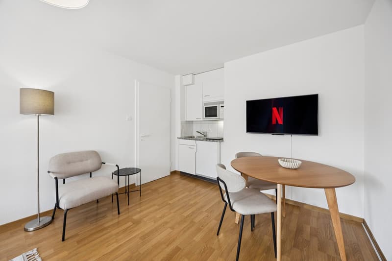 modern interior, white walls, hardwood floor, sofa, side table, dining table, chairs, lamp, flat screen TV, kitchen area with sink, faucet, and microwave, white cabinets