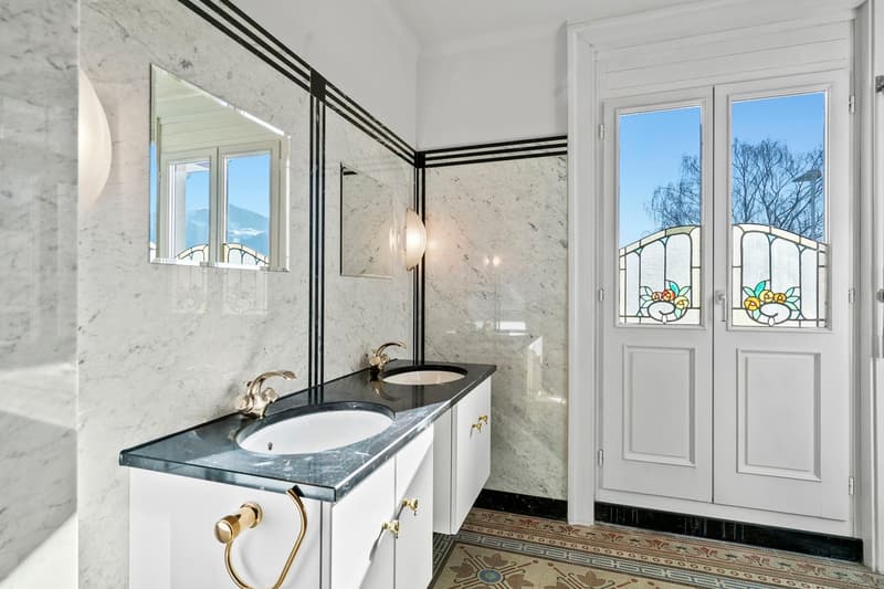 Spacious bathroom with marble walls, large vanity with marble countertop, gold fixtures, stained glass windows, and a view of the balcony/terrace outside