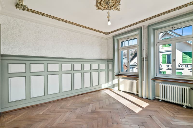 This image shows a spacious room with high ceilings, ornate moldings, and a large window overlooking the street. The walls feature a decorative paneled wainscoting in a light green color, and the floor is made of a herringbone pattern hardwood. The room h