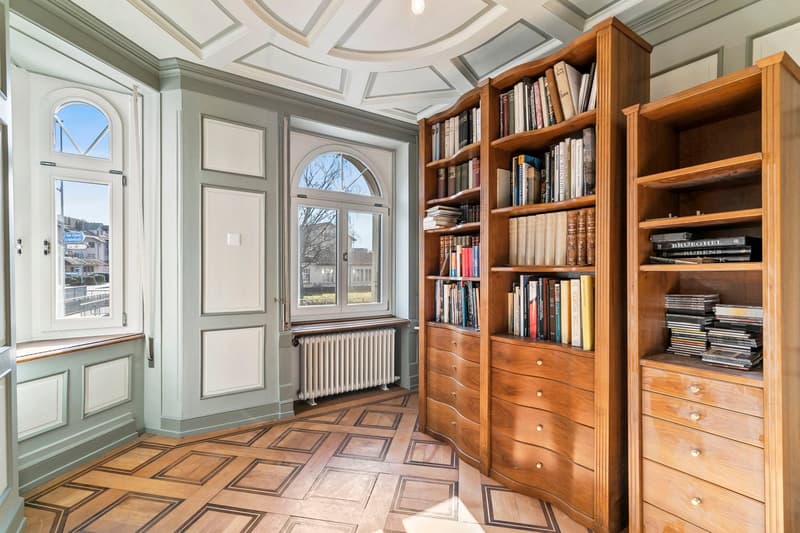 Spacious room with high ceilings, arched windows, and built-in wooden bookshelves and cabinets. The room has a decorative tiled floor and appears to be a study or library area.
