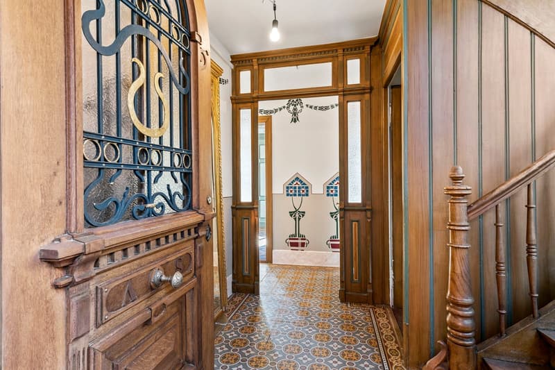 Ornate wooden entryway with intricate iron gate, decorative moldings, and patterned tile flooring. The space features a fireplace and leads to a hallway with more wooden paneling and decorative elements.