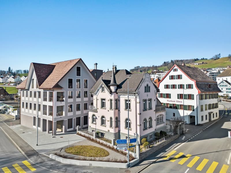 The image shows a picturesque town with a mix of traditional and modern buildings. The buildings have a variety of architectural styles, including some with balconies and terraces. The town appears to be situated in a hilly area with a clear blue sky in t