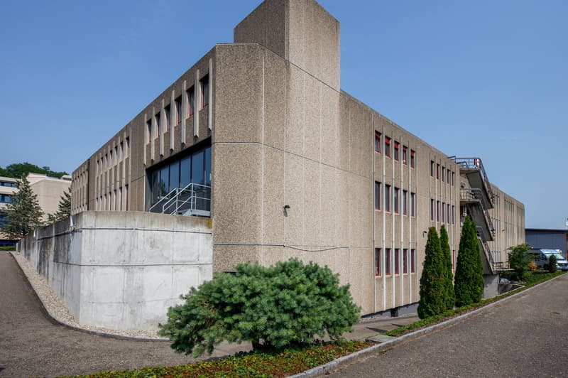 A large, multi-story concrete building with a distinctive architectural style. The building has a rectangular shape with a tower-like structure on one end. The exterior is made of concrete with a textured, granite-like finish. The building has several row
