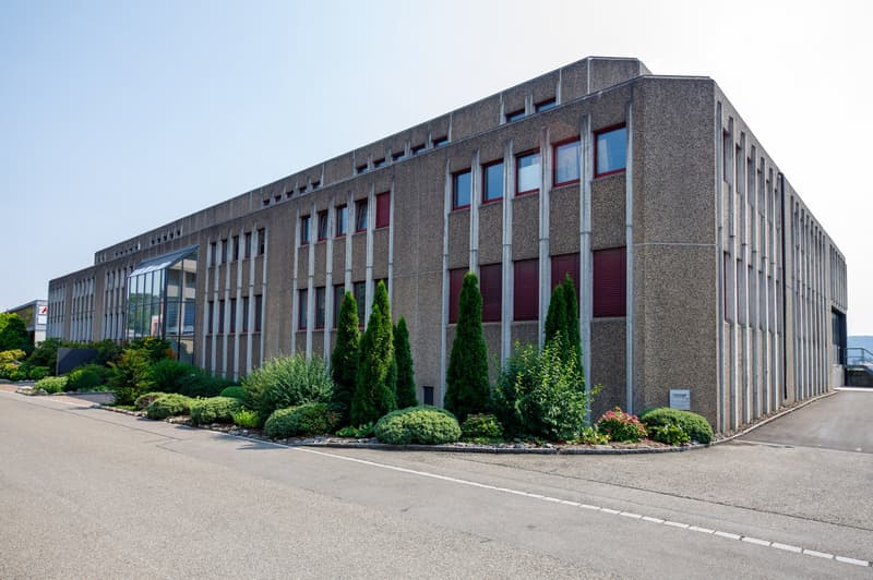 Large multi-story commercial building with a concrete exterior, red window frames, and landscaping with shrubs and trees around the perimeter. There appears to be a parking area in front of the building.