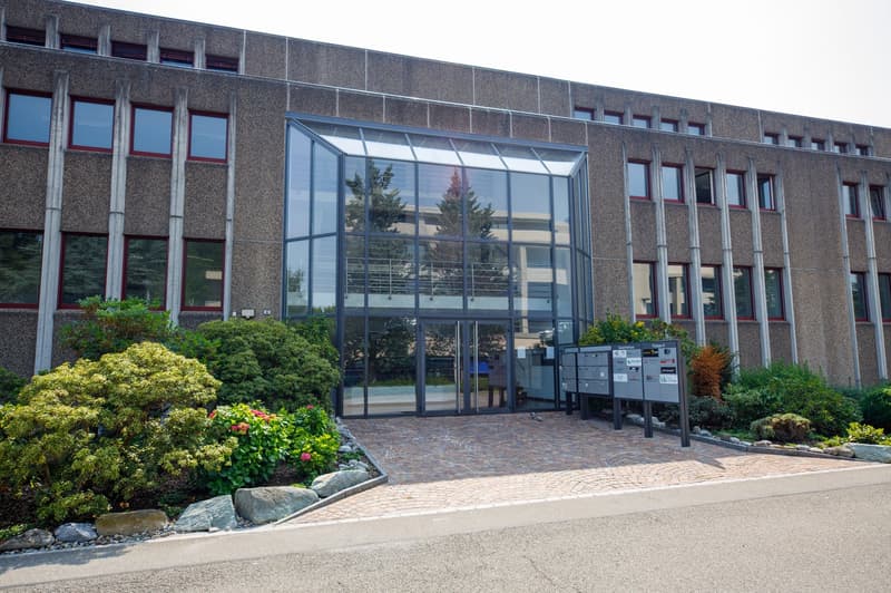Large multi-story office building with a glass entrance, surrounded by landscaping and a paved parking area