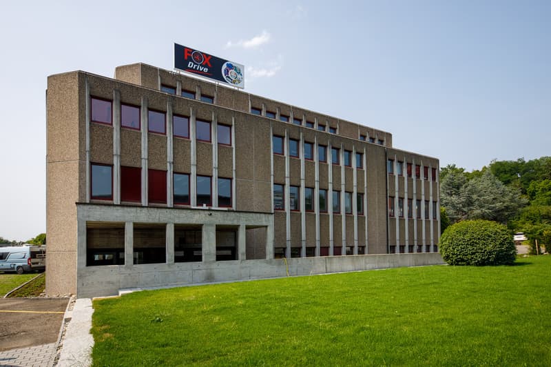 Multi-story concrete and glass office building with the Fox Drive logo on the top. The building has a rectangular shape with rows of windows and a grassy lawn in front.