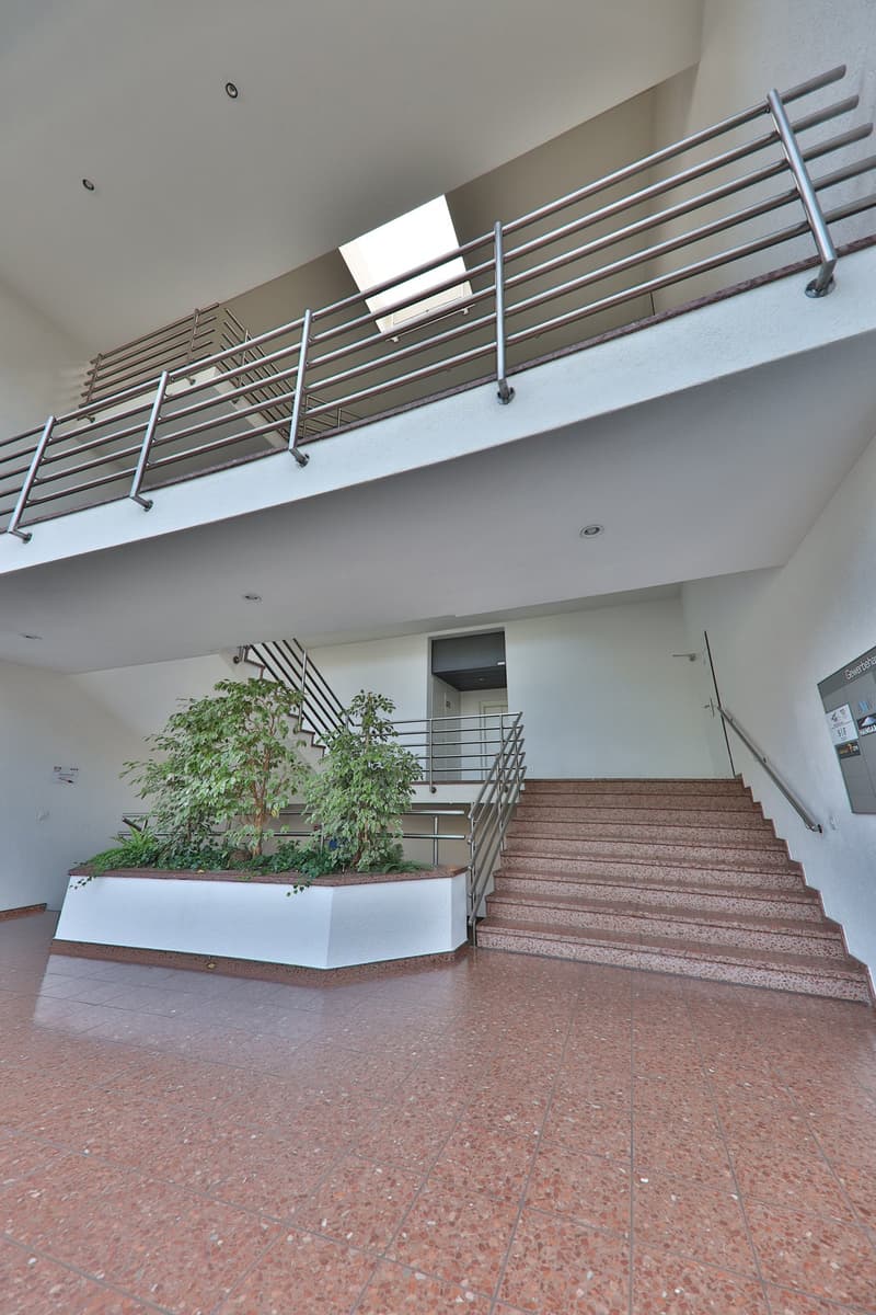 The image shows the entrance of a building with a staircase leading up to a balcony or terrace area. The stairs are made of granite or a similar stone material, and there are metal railings along the sides. There are also some potted plants placed at the