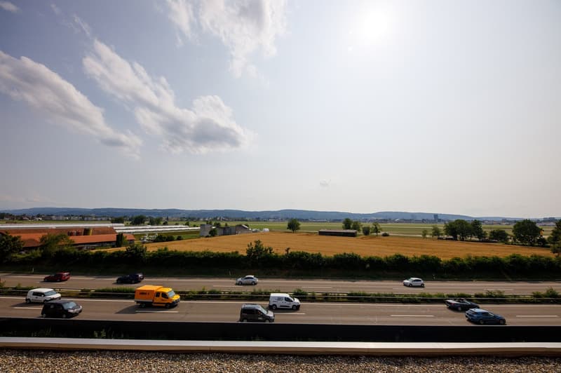 The image shows a scenic rural landscape with a highway running through it. There are fields of crops, trees, and buildings in the distance. The sky is partly cloudy with some white fluffy clouds. Several vehicles, including cars and a delivery van, can b
