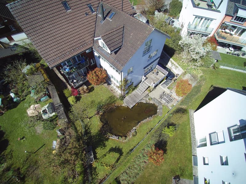 Aerial view of a house with a brown roof, white walls, a wooden deck, and a small pond in the garden.