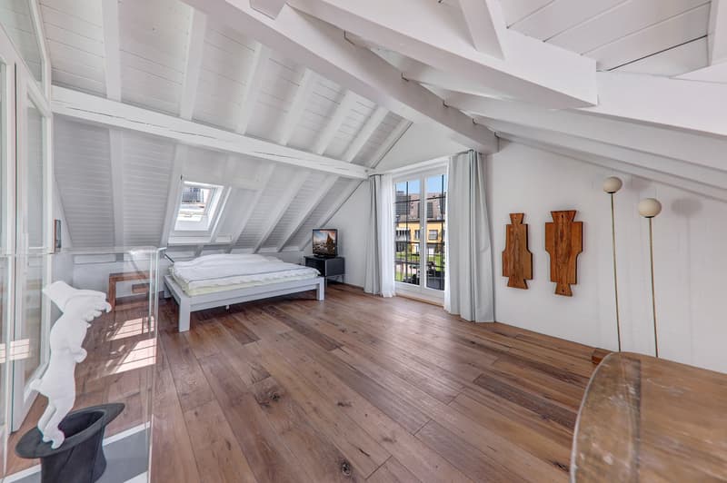 Spacious attic bedroom with white vaulted ceiling, hardwood floors, large windows with balcony access, and wooden decor elements