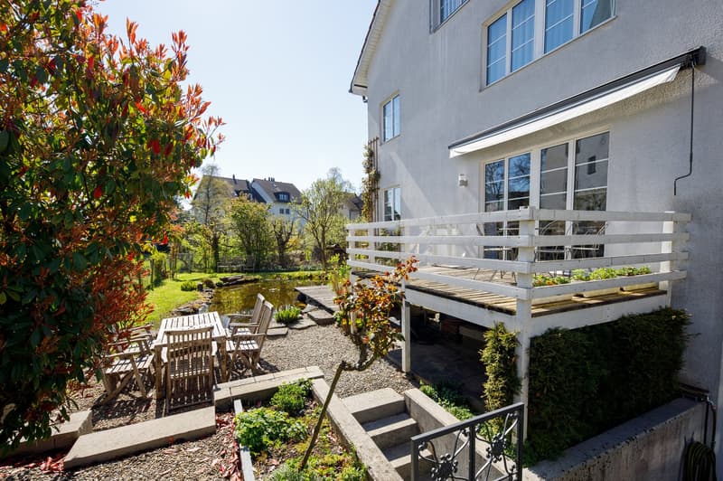A garden with a wooden deck, a table and chairs, a pond, and a white awning.