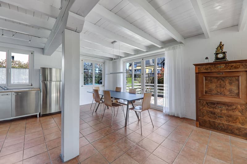 Dining room with a table and chairs, stainless steel kitchen appliances, wooden cabinet, tiled floor, white walls, white ceiling beams, glass doors, and windows.