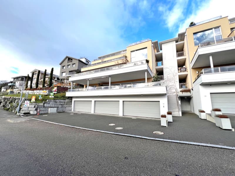 A residential building complex with two garages visible at the entrance, situated on a raised ground floor, and balconies visible on upper floors.