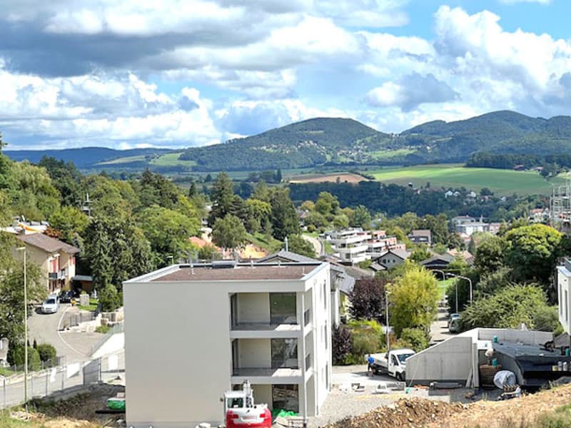 A white multi-story house under construction, with a few parked cars in front, on a sloping site with mountains and valleys in the background.