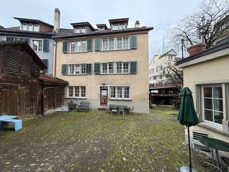 Two story residential building, beige facade, green shutters, a closed gate with railing, small paved courtyard with chairs, table, and an umbrella.