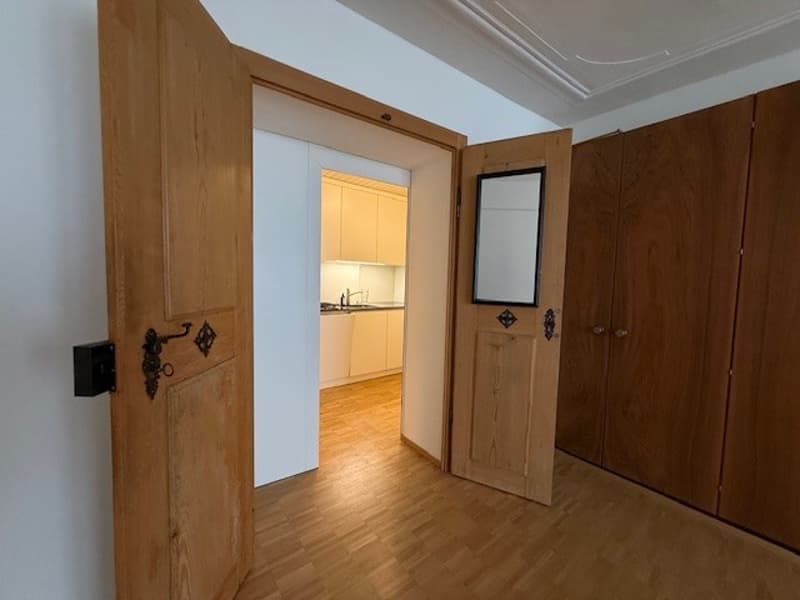 Light wood floor, two doors opening, white wall, empty room, kitchen at the end
