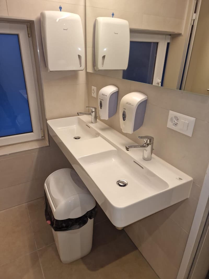 Bathroom with double sink, mirror, soap and paper towel dispensers, and electrical outlet.