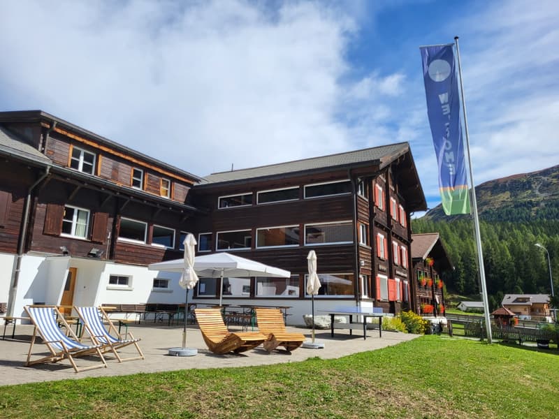 wooden building, multiple windows, patio with outdoor tables and chairs, tables, umbrellas, table tennis table, mountain in the background