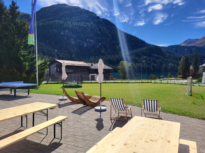 Outdoor area with tables and chairs, umbrellas, a tennis table, a wooden house in the background and a view of mountains.