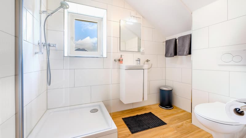 white tiled walls, white toilet, white shower, white sink, window, small countertop, toilet paper, shower mat, towels