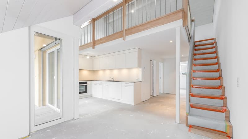 Open-concept living space with a kitchen, living room, and loft area. The kitchen features white cabinets and appliances. There is a sliding glass door leading to a balcony or terrace. The stairs leading to the loft area have a modern, wooden design.