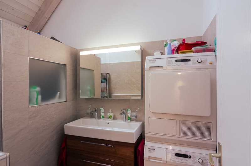 small white vanity, modern mirror, wooden cabinet, under cabinet drawers, large white washing machine and dryer, white tiled walls, ceiling lights