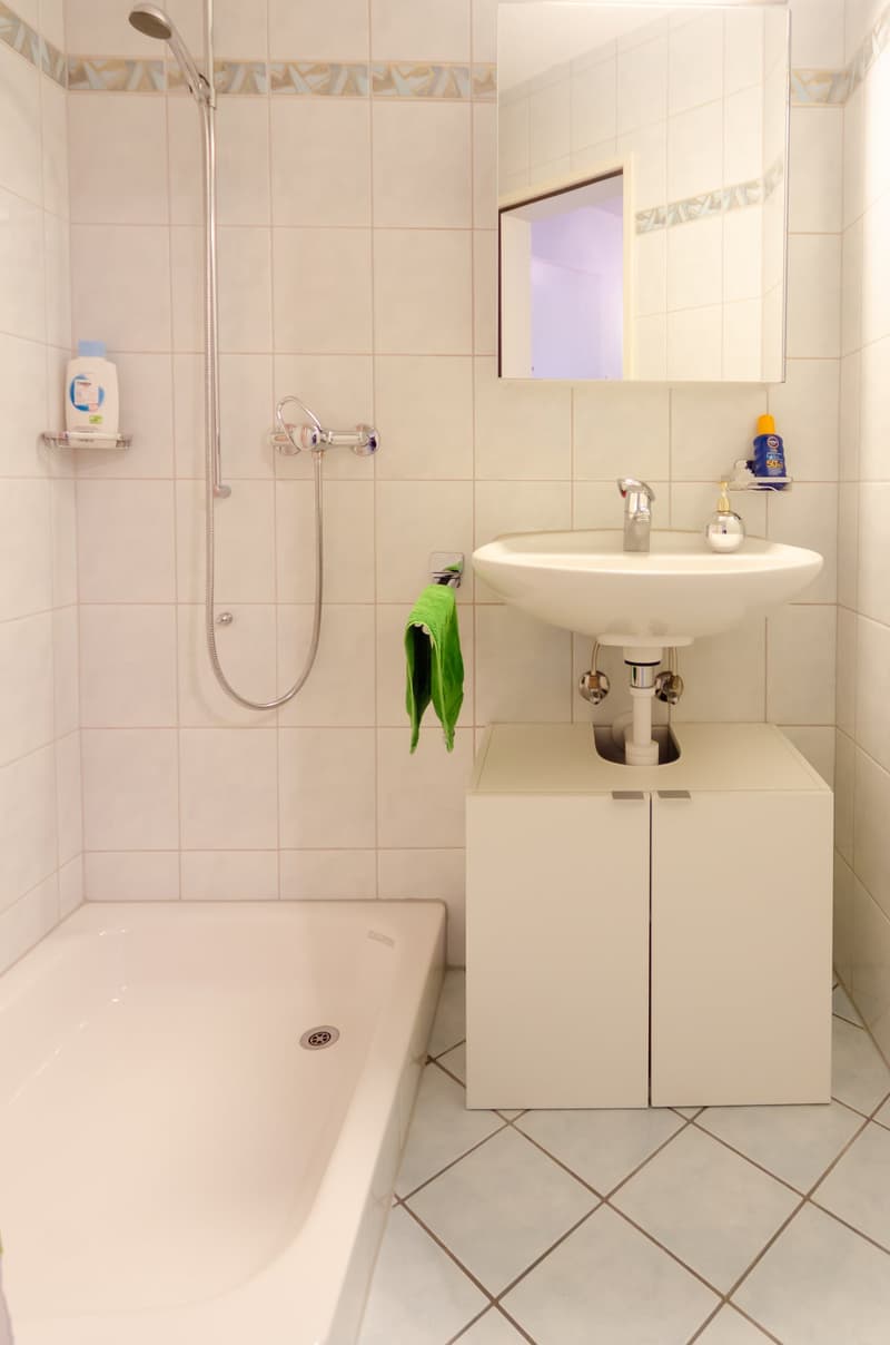 A small bathroom with a bathtub, sink, mirror, and a cabinet with a shower head and hose. Tiled floors, white walls, and a single white sink.