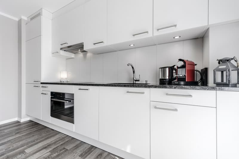 Modern, minimalist kitchen with white cabinets, black granite countertops, built-in oven, and small appliances such as a coffee maker and kettle on the counter