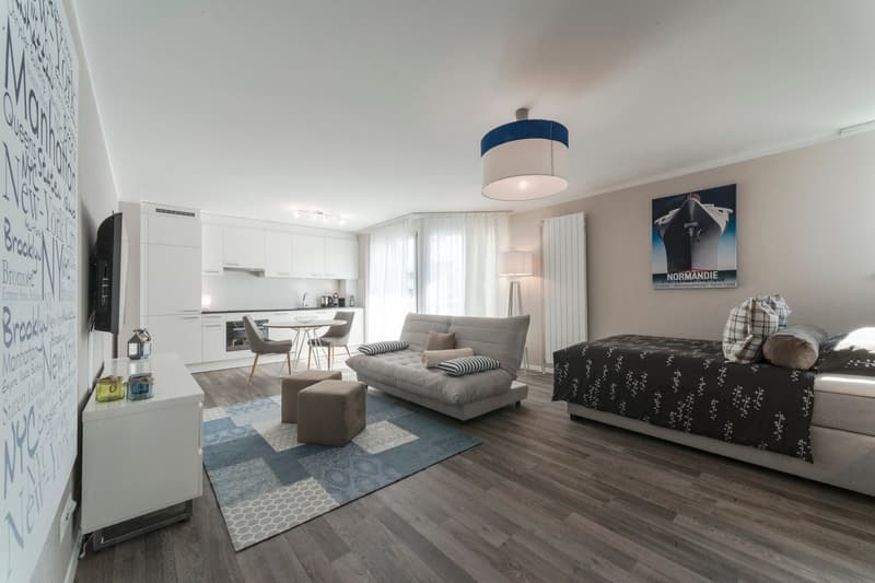 Open-concept studio apartment with hardwood floors, white walls, large windows, living area with sofa and coffee table, kitchen with white cabinets and appliances, and a bed area with a patterned bedspread.