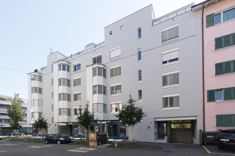 Multi-story apartment building with gray exterior, large windows, and balconies. There is a parking area in front of the building.