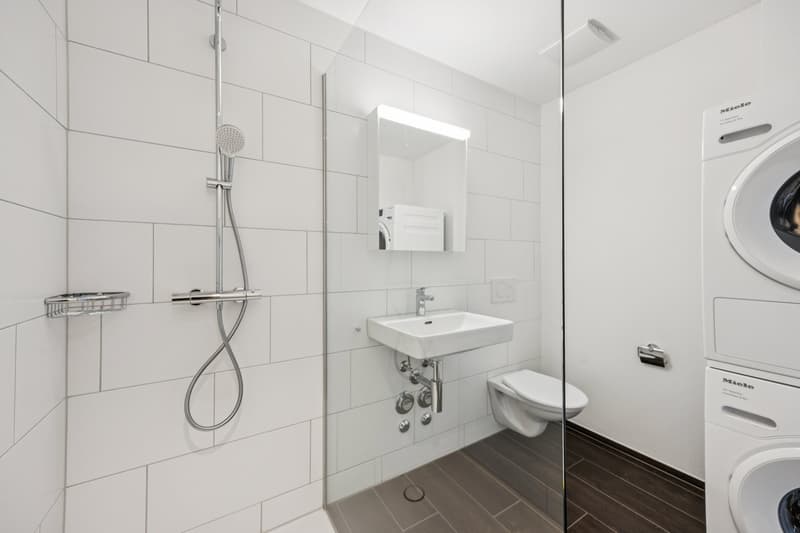 modern white tiled bathroom with a sink, toilet, shower, and a washing machine