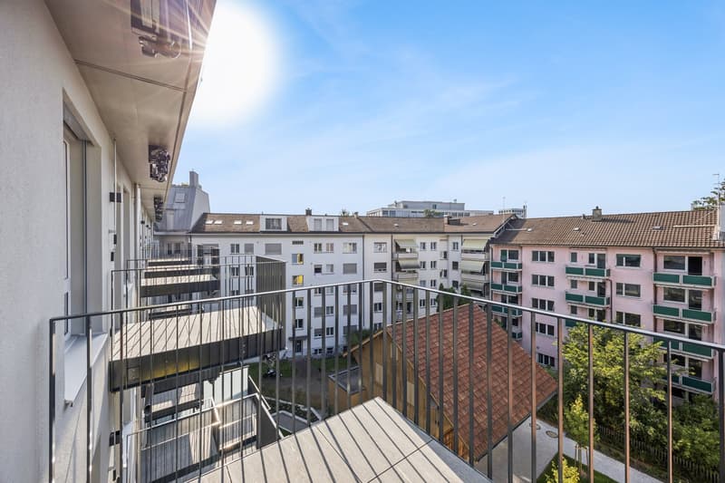 Modern building with multiple floors, terrace with metal railing, balcony, multiple buildings in the background, green trees, and buildings.