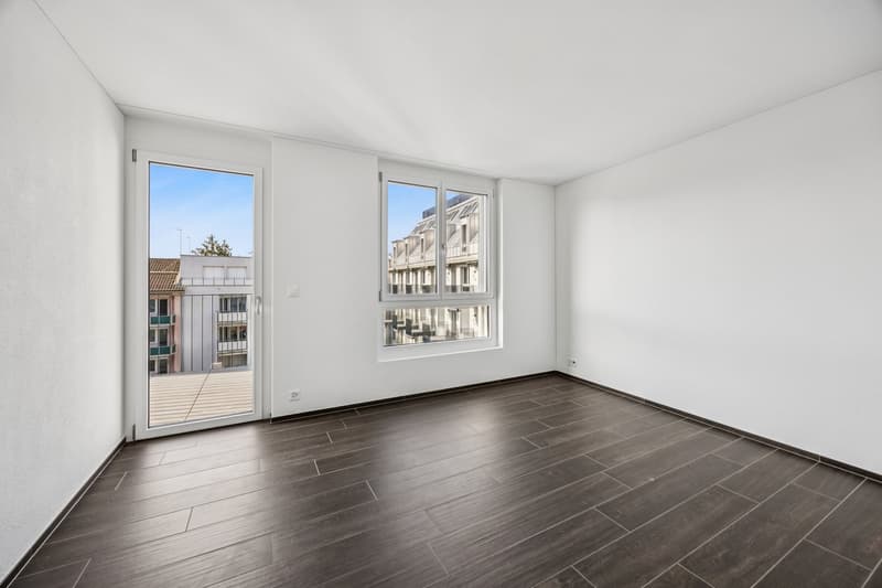 An empty room with white walls, wooden floor, sliding door leading to a balcony, and windows