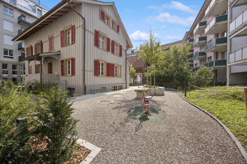 Two-story house with red shutters, wooden entrance, gravel path, child's play area, nearby apartment building