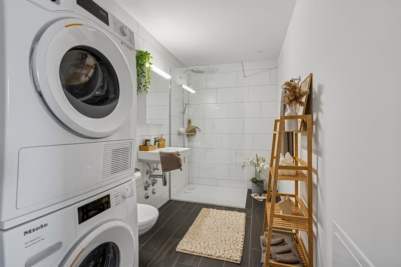 Bathroom with white tiles, a shower, toilet, sink, and a wooden shelf with towels and toiletries. Next to the bathroom is a washer dryer.