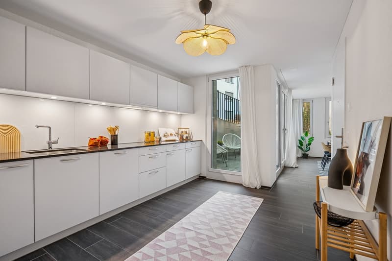 Modern kitchen with white cabinets, black countertops, modern pendant lighting, white curtains, wooden rug, and access to a balcony.