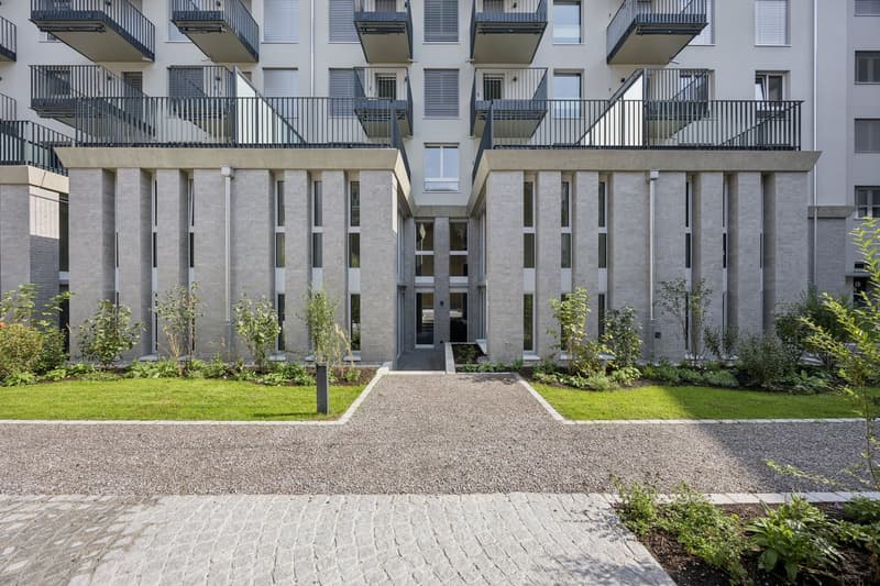 white building, grey exterior, multiple balconies, brick facade, green entrance pathway, planted garden, concrete paths