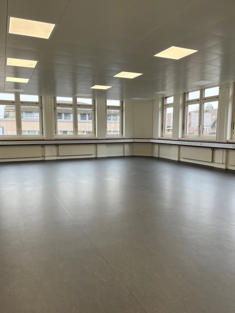 Large open room with white walls, recessed lighting, and large windows. The room has a polished concrete floor and appears to be a multipurpose space, possibly a dance studio or community center.