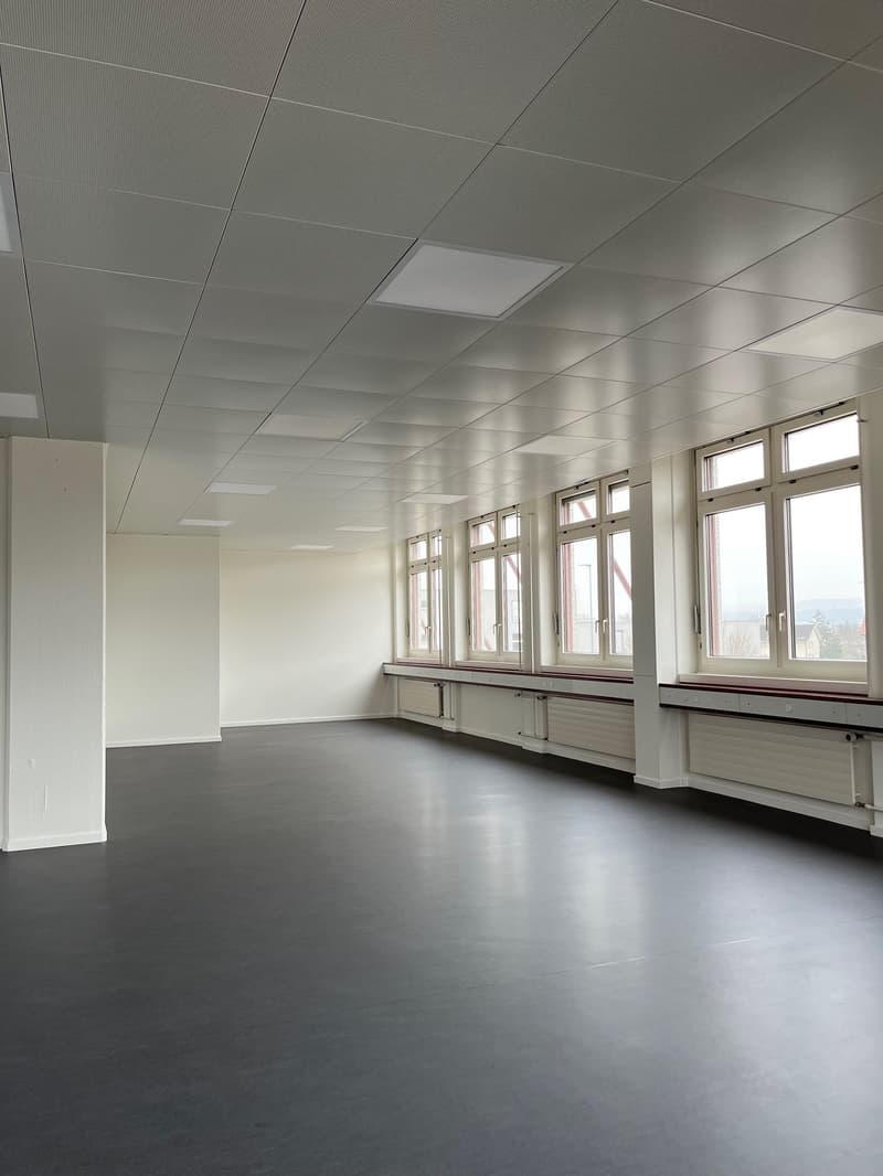 The image shows a large, open office space with a high ceiling and large windows along one wall. The room has a clean, modern design with white walls and a gray tiled floor. The ceiling is covered with a grid of recessed lighting panels. The space appears