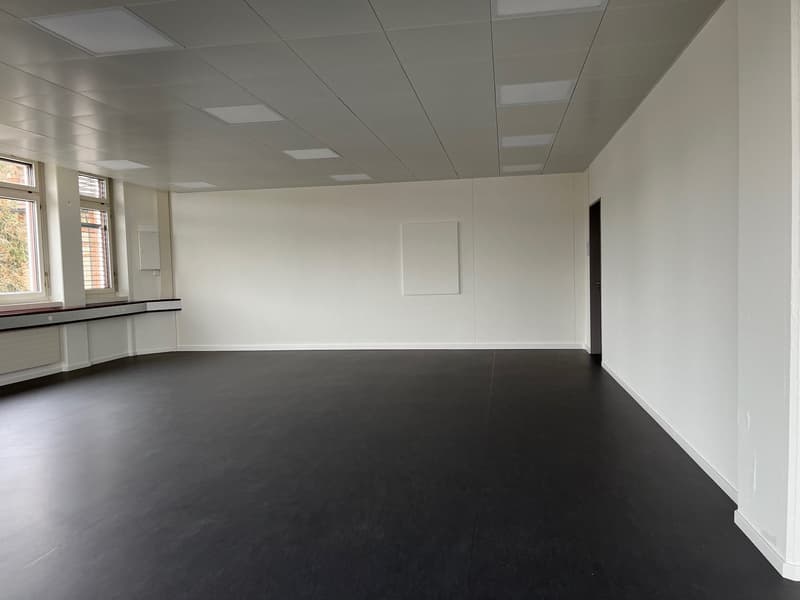 The image shows a large, open office space with white walls, a tiled ceiling, and a dark gray floor. The room appears to be empty, with no furniture or other objects visible. The space has several windows along one wall, providing natural light. Overall,