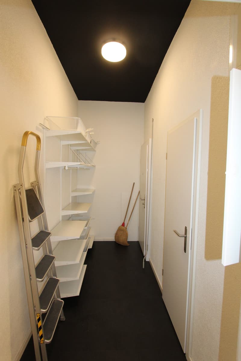 A hallway with white walls, black floor, white shelves, a ladder, and a broom