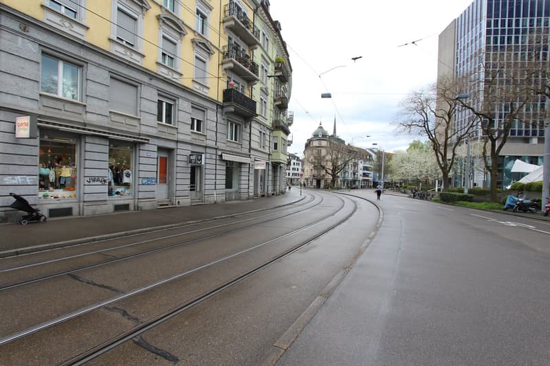 Empty street with tram tracks, buildings on both sides, shops, trees, and parked bicycles.